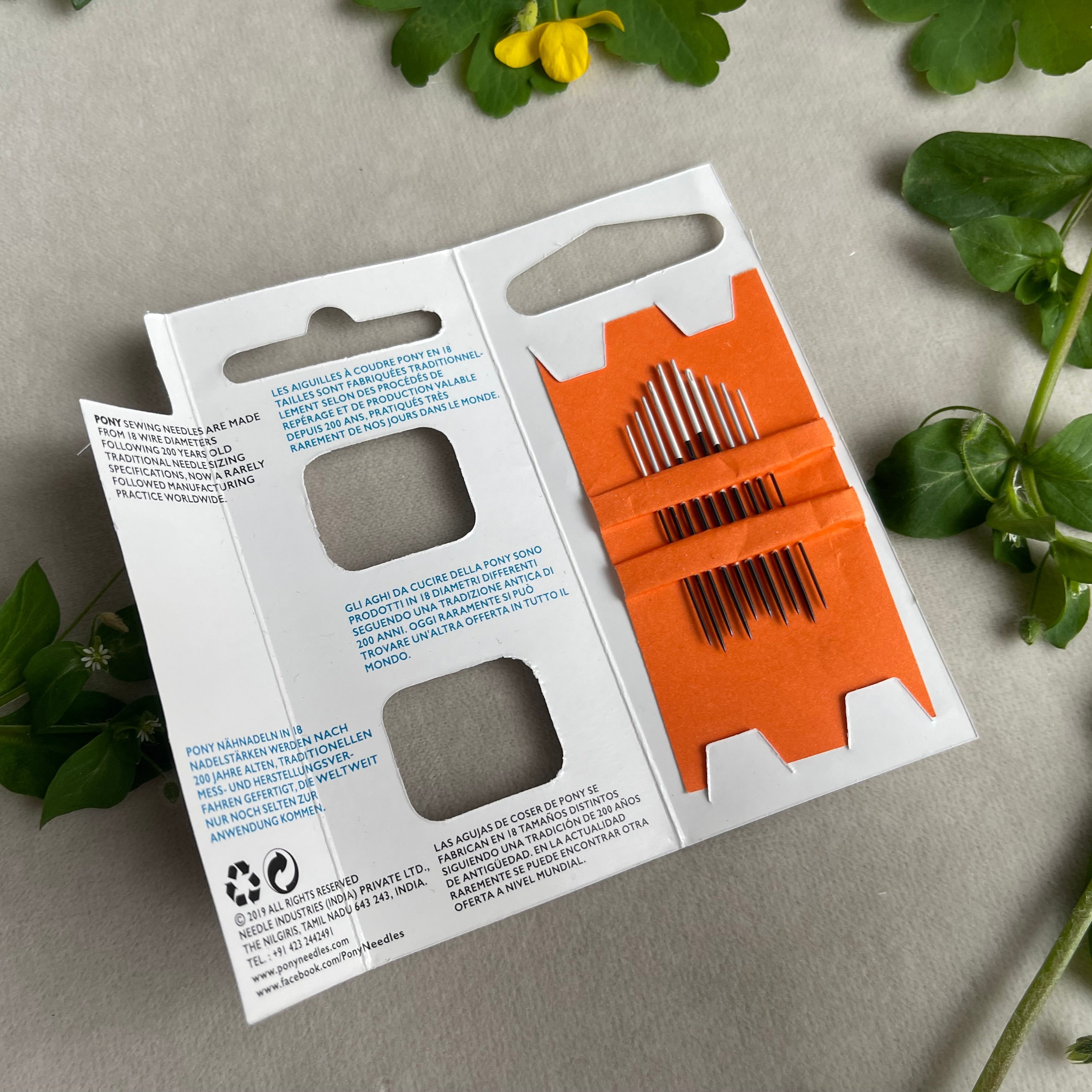 Packaging of a sewing kit with orange components on a gray surface with green leaves.