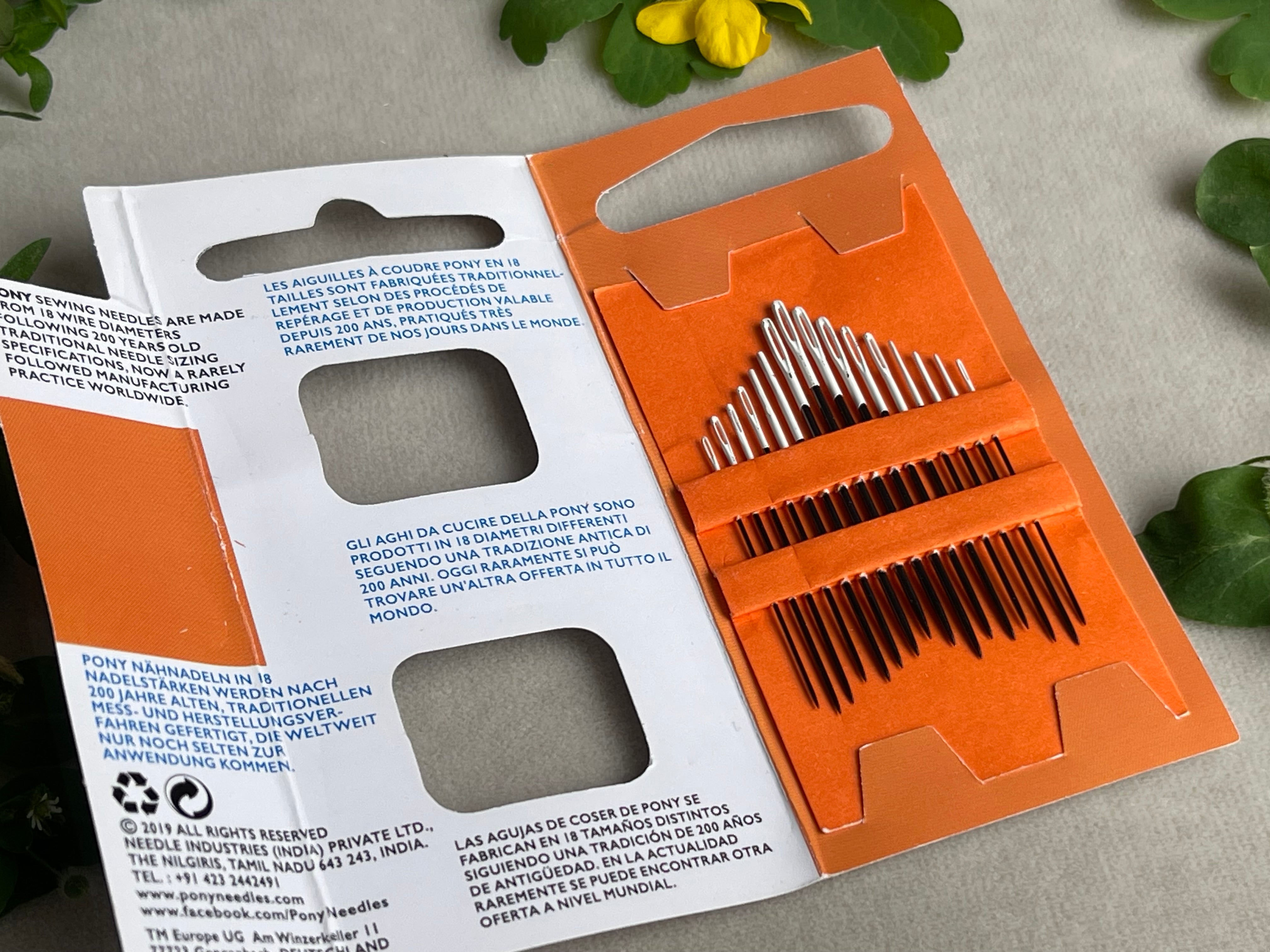 Set of needles in an orange and white packaging on a light gray background with green leaves.